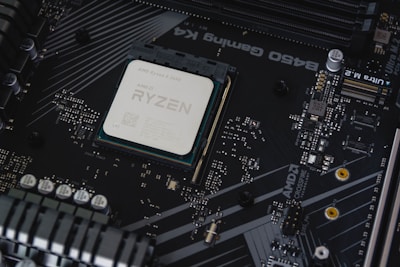 A computer motherboard with a prominently visible AMD Ryzen processor installed. Circuits, transistors, and various electronic components are integrated into the board. The background features a dark, matte texture with intricate patterns and electronic paths.