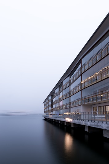 A modern, multi-story building with large glass windows extends along a waterfront, with calm water reflecting the building