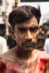 A man with visible red stains on his face and body stands in a crowd with a serious expression. His hair is unkempt and he appears to be in an urban setting with blurred figures in the background.