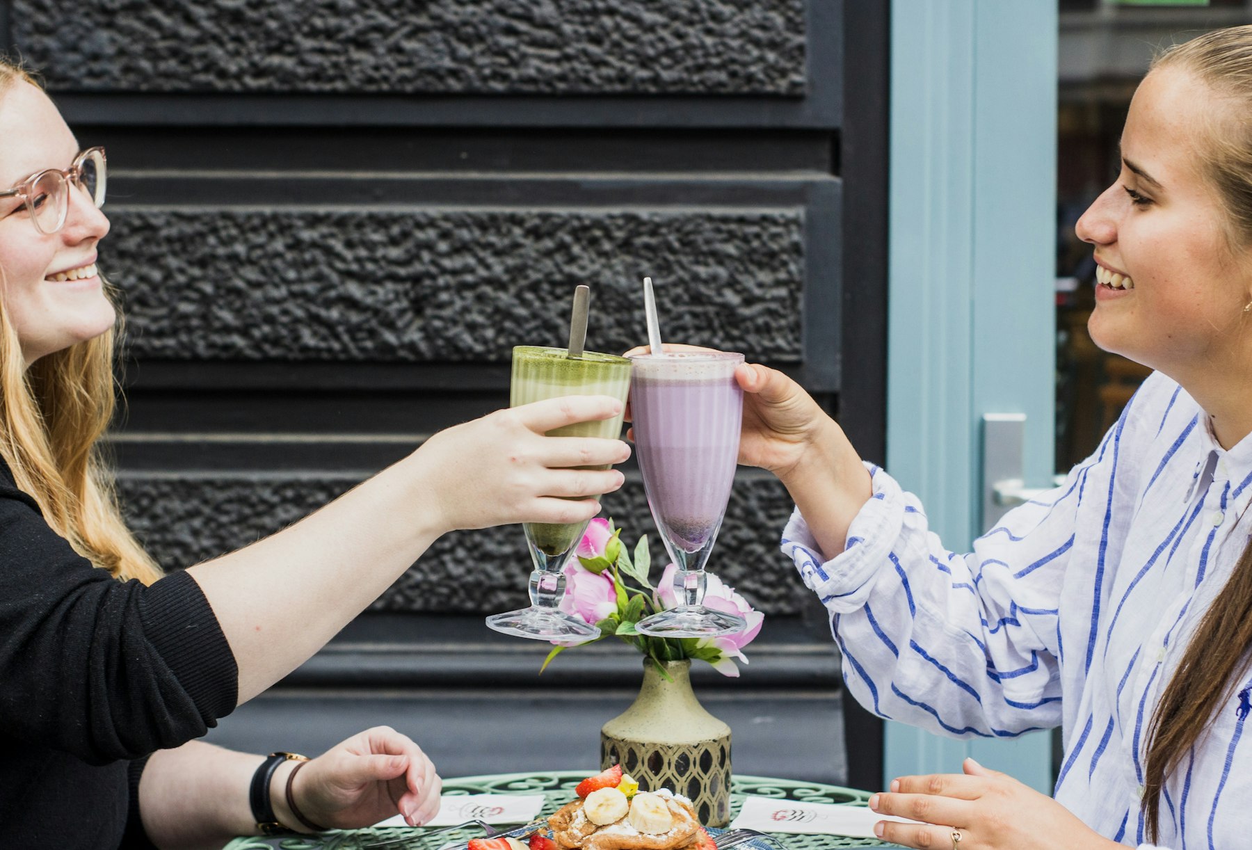 Two friends toasting fresh smoothies at a sidewalk table