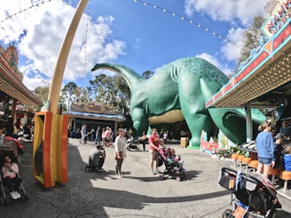 A lively amusement park scene featuring a large green dinosaur statue surrounded by colorful buildings and decorations. Families with strollers and children are scattered around, enjoying the pleasant day. The sky is bright with soft clouds, adding to the cheerful atmosphere.