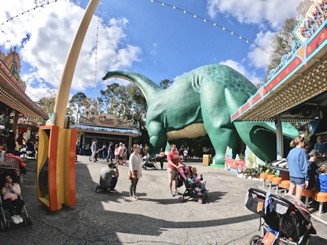 A lively amusement park scene featuring a large green dinosaur statue surrounded by colorful buildings and decorations. Families with strollers and children are scattered around, enjoying the pleasant day. The sky is bright with soft clouds, adding to the cheerful atmosphere.