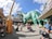 A lively amusement park scene featuring a large green dinosaur statue surrounded by colorful buildings and decorations. Families with strollers and children are scattered around, enjoying the pleasant day. The sky is bright with soft clouds, adding to the cheerful atmosphere.