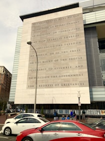 A large building facade with the text of the First Amendment of the United States Constitution engraved prominently. Several cars, including a red taxi, are parked in front of the building. People are sitting and standing near the entrance, and a lamppost is visible in the foreground.