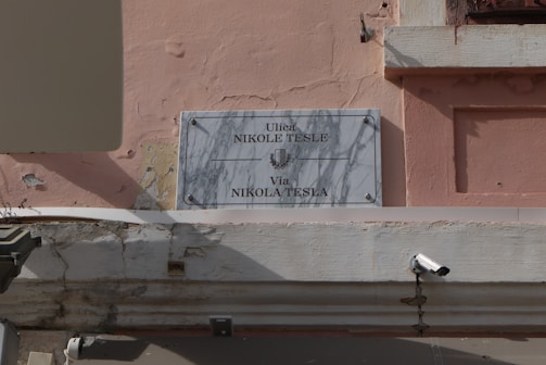 A street sign on a textured peach-colored wall with white marble showing the names Ulica Nikole Tesle and Via Nikola Tesla. The sign is held in place by metal bolts and a surveillance camera is mounted on the wall nearby.
