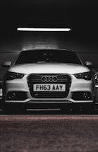 A white car with a prominently displayed license plate 'FH63 AAY' is parked in a dimly lit indoor space. The lighting highlights the sleek design and logo on the grille.
