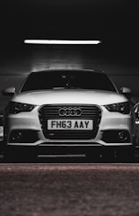 A white car with a prominently displayed license plate 'FH63 AAY' is parked in a dimly lit indoor space. The lighting highlights the sleek design and logo on the grille.