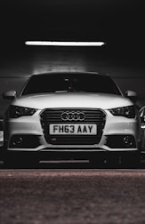 A white car with a prominently displayed license plate 'FH63 AAY' is parked in a dimly lit indoor space. The lighting highlights the sleek design and logo on the grille.