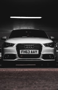 A white car with a prominently displayed license plate 'FH63 AAY' is parked in a dimly lit indoor space. The lighting highlights the sleek design and logo on the grille.