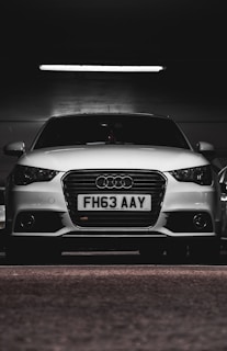A white car with a prominently displayed license plate 'FH63 AAY' is parked in a dimly lit indoor space. The lighting highlights the sleek design and logo on the grille.