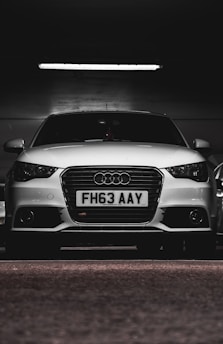 A white car with a prominently displayed license plate 'FH63 AAY' is parked in a dimly lit indoor space. The lighting highlights the sleek design and logo on the grille.