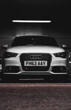 A white car with a prominently displayed license plate 'FH63 AAY' is parked in a dimly lit indoor space. The lighting highlights the sleek design and logo on the grille.