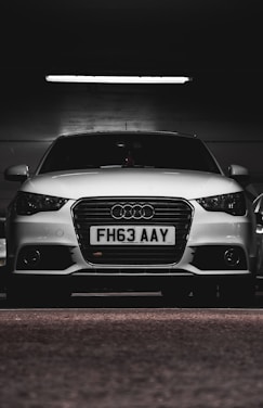 A white car with a prominently displayed license plate 'FH63 AAY' is parked in a dimly lit indoor space. The lighting highlights the sleek design and logo on the grille.