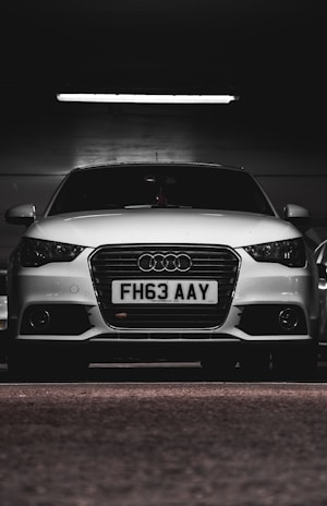 A white car with a prominently displayed license plate 'FH63 AAY' is parked in a dimly lit indoor space. The lighting highlights the sleek design and logo on the grille.
