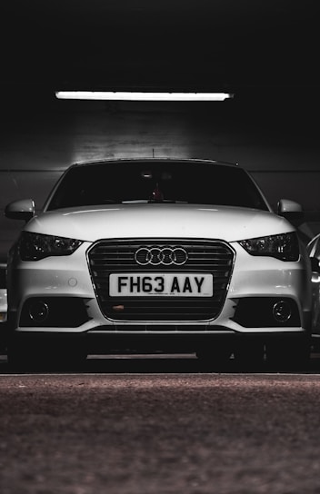 A white car with a prominently displayed license plate 'FH63 AAY' is parked in a dimly lit indoor space. The lighting highlights the sleek design and logo on the grille.