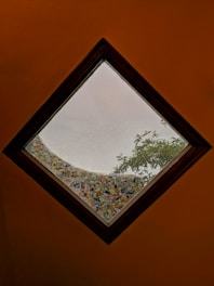 A skylight with a diamond shape provides a view to the outside, showing a mosaic design around the edge of the window. There is a tree branch visible through the glass, and the sky appears overcast. The frame of the skylight is made from a dark wood.