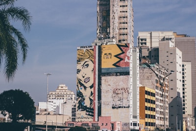 A large cityscape with tall buildings featuring a prominent mural on one of the structures. The mural displays pop-art style with a comic illustration of a woman's face and vibrant geometric shapes. The buildings have a mix of modern and older architectural styles, with some graffiti and signs of weathering.