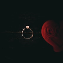 A delicate gold ring with a small diamond is placed against a dark background. On the right side, a blurred red rose enhances the romantic feel.