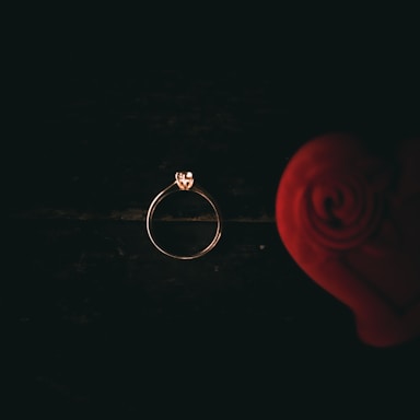 A delicate gold ring with a small diamond is placed against a dark background. On the right side, a blurred red rose enhances the romantic feel.