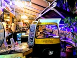 A cozy, dimly lit indoor area with eclectic decor featuring a repurposed auto rickshaw. A blackboard displays happy hour specials and a variety of seating options are available, including stool bars and cushioned benches. Soft lighting and plants create a welcoming ambiance.