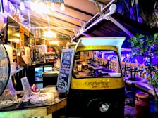 A cozy, dimly lit indoor area with eclectic decor featuring a repurposed auto rickshaw. A blackboard displays happy hour specials and a variety of seating options are available, including stool bars and cushioned benches. Soft lighting and plants create a welcoming ambiance.