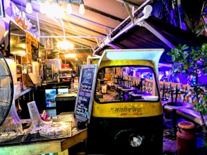 A cozy, dimly lit indoor area with eclectic decor featuring a repurposed auto rickshaw. A blackboard displays happy hour specials and a variety of seating options are available, including stool bars and cushioned benches. Soft lighting and plants create a welcoming ambiance.
