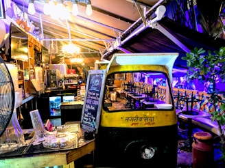 A cozy, dimly lit indoor area with eclectic decor featuring a repurposed auto rickshaw. A blackboard displays happy hour specials and a variety of seating options are available, including stool bars and cushioned benches. Soft lighting and plants create a welcoming ambiance.