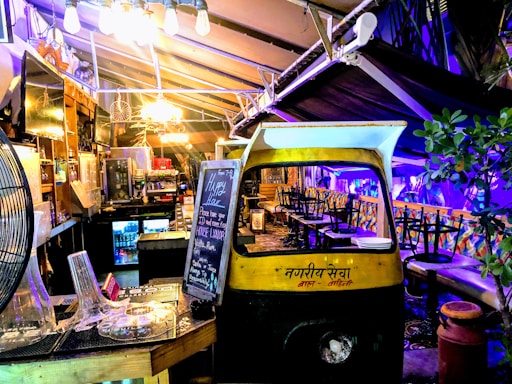 A cozy, dimly lit indoor area with eclectic decor featuring a repurposed auto rickshaw. A blackboard displays happy hour specials and a variety of seating options are available, including stool bars and cushioned benches. Soft lighting and plants create a welcoming ambiance.