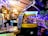 A cozy, dimly lit indoor area with eclectic decor featuring a repurposed auto rickshaw. A blackboard displays happy hour specials and a variety of seating options are available, including stool bars and cushioned benches. Soft lighting and plants create a welcoming ambiance.