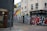 A street corner featuring vibrant urban graffiti on the walls of several buildings, including a colorful mural on the side of a building with abstract geometric shapes in yellow, blue, and red. One building is painted dark gray, while another has a cream facade adorned with graffiti. A person carrying a bag walks past a shuttered storefront with large bold graffiti. Signs above indicate the presence of a bistrot and a fondue bar.