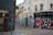 A street corner featuring vibrant urban graffiti on the walls of several buildings, including a colorful mural on the side of a building with abstract geometric shapes in yellow, blue, and red. One building is painted dark gray, while another has a cream facade adorned with graffiti. A person carrying a bag walks past a shuttered storefront with large bold graffiti. Signs above indicate the presence of a bistrot and a fondue bar.