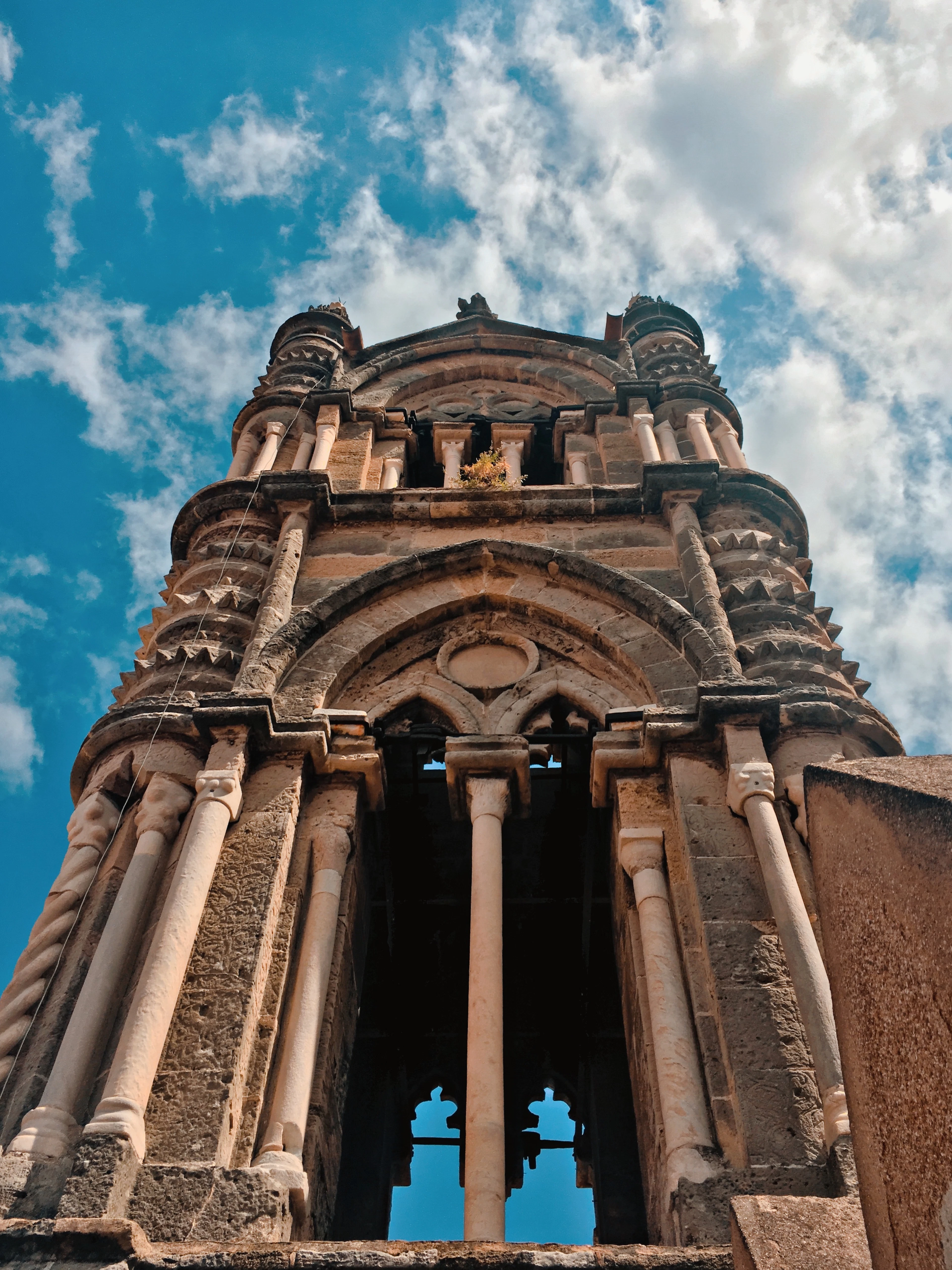 Looking up at a carved stone bell tower of Palermo Cathedral against a blue sky streaked with clouds