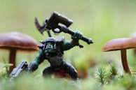 A small, detailed figurine resembling an orc-like creature is posed in an action stance with a large axe raised above its head. The creature has green skin, wears armor, and stands among natural elements like moss and small mushrooms. The background is blurred, providing a miniature forest setting.