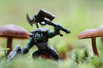 A small, detailed figurine resembling an orc-like creature is posed in an action stance with a large axe raised above its head. The creature has green skin, wears armor, and stands among natural elements like moss and small mushrooms. The background is blurred, providing a miniature forest setting.