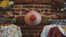 A whimsical setting with a large plush pig head mounted on a brick wall, flanked by various animal-themed decor. Below, two clothing racks display children's clothes with playful designs, emphasizing a cozy and imaginative atmosphere.