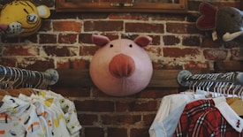 A whimsical setting with a large plush pig head mounted on a brick wall, flanked by various animal-themed decor. Below, two clothing racks display children's clothes with playful designs, emphasizing a cozy and imaginative atmosphere.