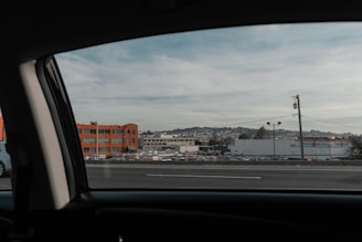 The image captures a view from inside a vehicle looking at an urban landscape. It features a large orange building, a warehouse with 'TRUCK' signage, and a distant view of hills with residential areas. Power lines and several parked vehicles are visible in the foreground.