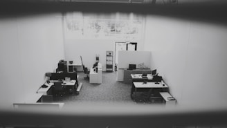 An office space is captured from a high angle, showing multiple desks equipped with computers, chairs, and office supplies. The space is well-lit due to a large window providing natural light. The walls are plain and the floor is carpeted. The environment appears tidy and sparsely populated.