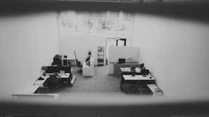 An office space is captured from a high angle, showing multiple desks equipped with computers, chairs, and office supplies. The space is well-lit due to a large window providing natural light. The walls are plain and the floor is carpeted. The environment appears tidy and sparsely populated.