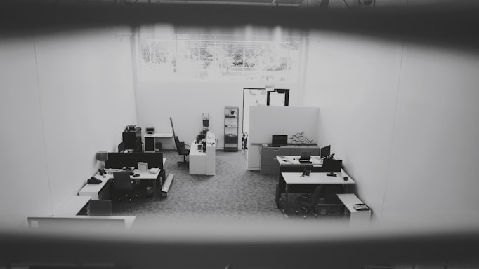 An office space is captured from a high angle, showing multiple desks equipped with computers, chairs, and office supplies. The space is well-lit due to a large window providing natural light. The walls are plain and the floor is carpeted. The environment appears tidy and sparsely populated.