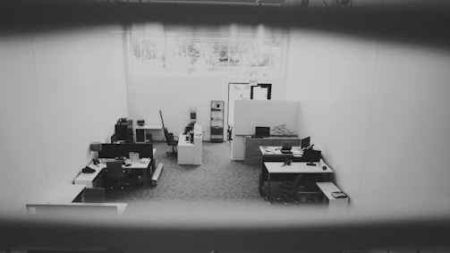 An office space is captured from a high angle, showing multiple desks equipped with computers, chairs, and office supplies. The space is well-lit due to a large window providing natural light. The walls are plain and the floor is carpeted. The environment appears tidy and sparsely populated.