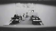 An office space is captured from a high angle, showing multiple desks equipped with computers, chairs, and office supplies. The space is well-lit due to a large window providing natural light. The walls are plain and the floor is carpeted. The environment appears tidy and sparsely populated.