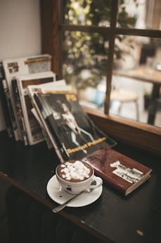 A cozy corner in a room featuring a stack of magazines and a hardcover book on a dark wooden surface beside a window. A cup of hot chocolate with marshmallows sits on a saucer with a spoon. The scene is softly lit with natural light coming through the window, giving a warm and inviting ambiance.