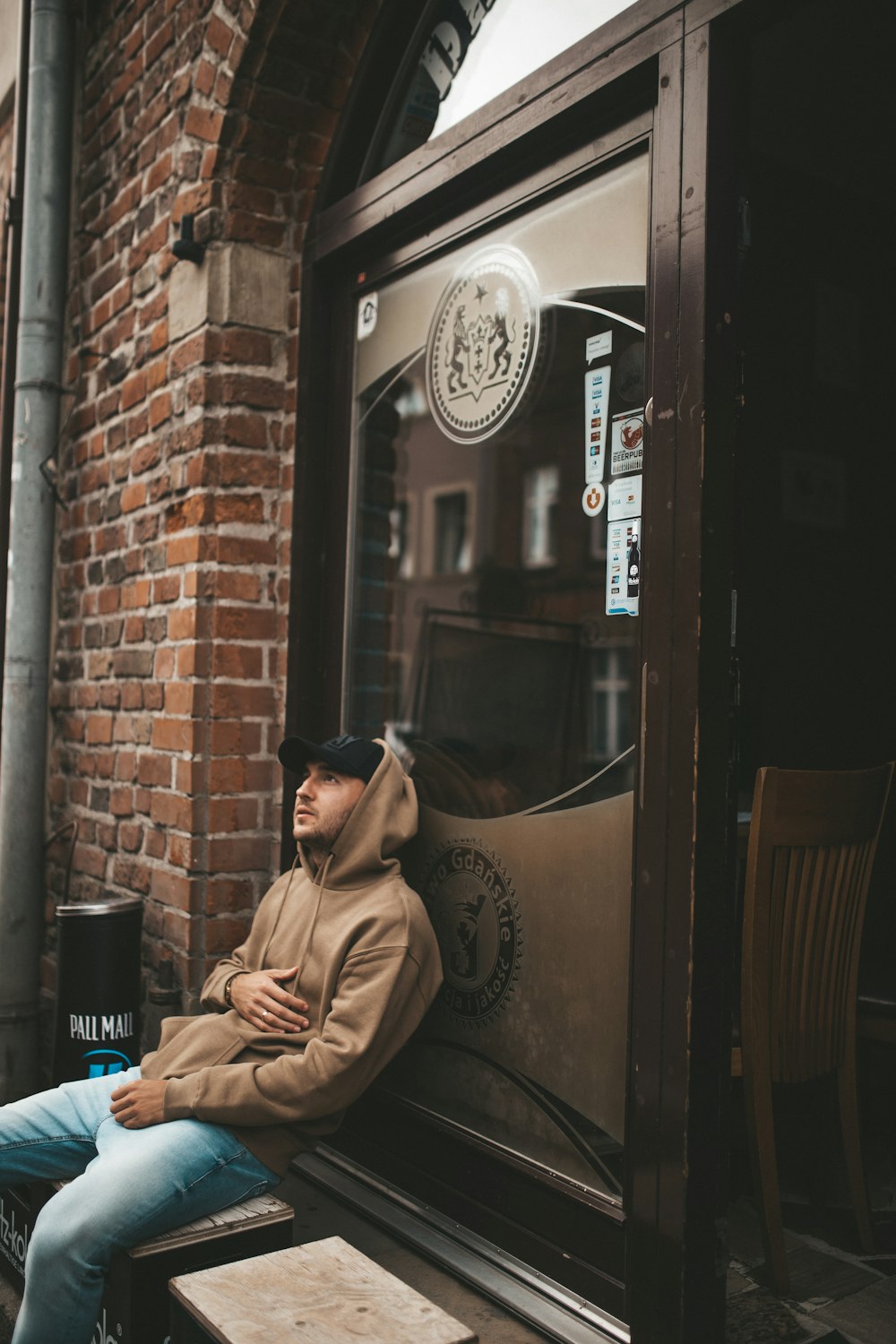 man wearing brown pullover hoodie leaning on glass wall looking up during daytime