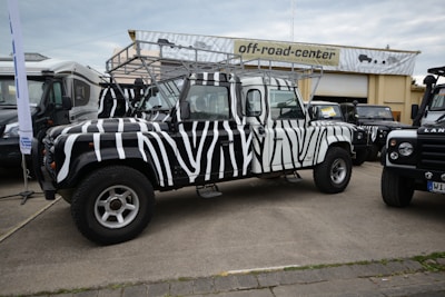 A zebra-striped off-road vehicle is parked in front of a building with a sign that reads 'off-road-center'. The vehicle has distinctive black and white stripes, large tires, and a roof rack. Other vehicles are parked nearby, including another Land Rover, all on a paved area.