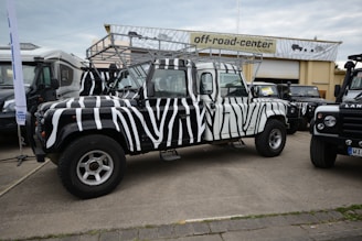 A zebra-striped off-road vehicle is parked in front of a building with a sign that reads 'off-road-center'. The vehicle has distinctive black and white stripes, large tires, and a roof rack. Other vehicles are parked nearby, including another Land Rover, all on a paved area.