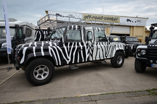 A zebra-striped off-road vehicle is parked in front of a building with a sign that reads 'off-road-center'. The vehicle has distinctive black and white stripes, large tires, and a roof rack. Other vehicles are parked nearby, including another Land Rover, all on a paved area.