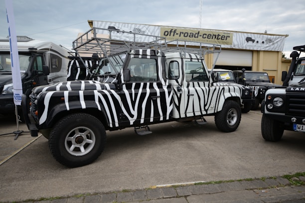 A zebra-striped off-road vehicle is parked in front of a building with a sign that reads 'off-road-center'. The vehicle has distinctive black and white stripes, large tires, and a roof rack. Other vehicles are parked nearby, including another Land Rover, all on a paved area.