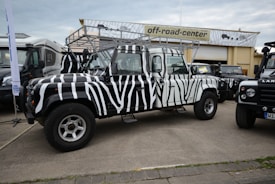 A zebra-striped off-road vehicle is parked in front of a building with a sign that reads 'off-road-center'. The vehicle has distinctive black and white stripes, large tires, and a roof rack. Other vehicles are parked nearby, including another Land Rover, all on a paved area.