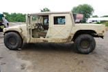 A rugged off-road vehicle with a beige exterior, covered in mud, is parked on a dirt path. The front door on the driver's side is open, revealing a simple interior with a steering wheel and seats. The vehicle has large, thick tires suitable for off-road terrain. In the background, there are some trees and a signboard, along with a cloudy sky.